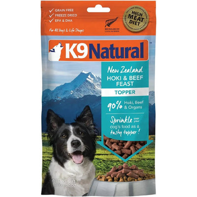 A bag of K9 Natural Hoki & Beef Grain-Free Freeze-Dried Dog Food Topper displays a happy black and white dog, mountain scenery, and product details such as “90% Hoki, Beef & Organs” and “Grain Free.”.